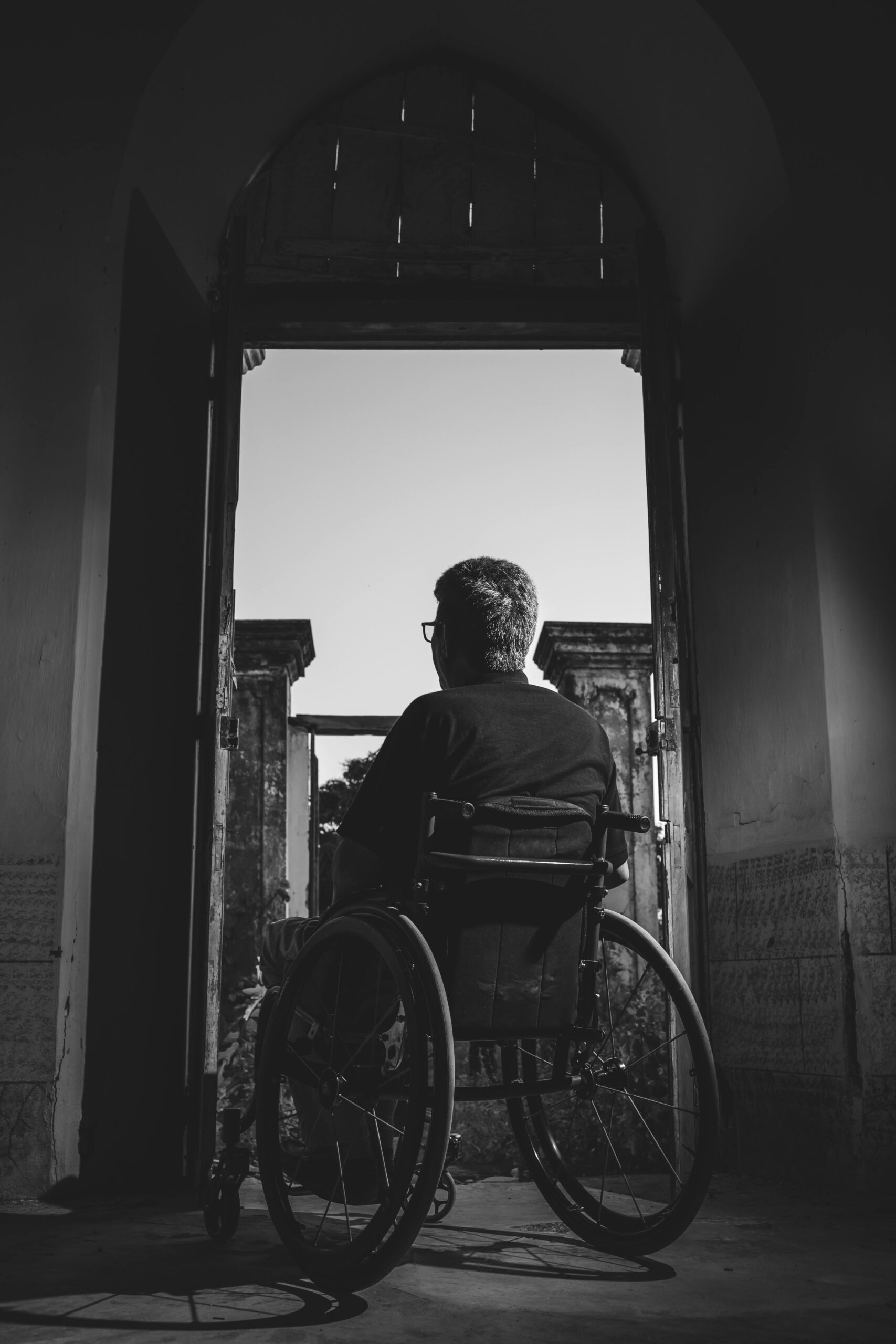 Black and white photo of a man in a wheelchair looking outside through an open doorway, evoking solitude.