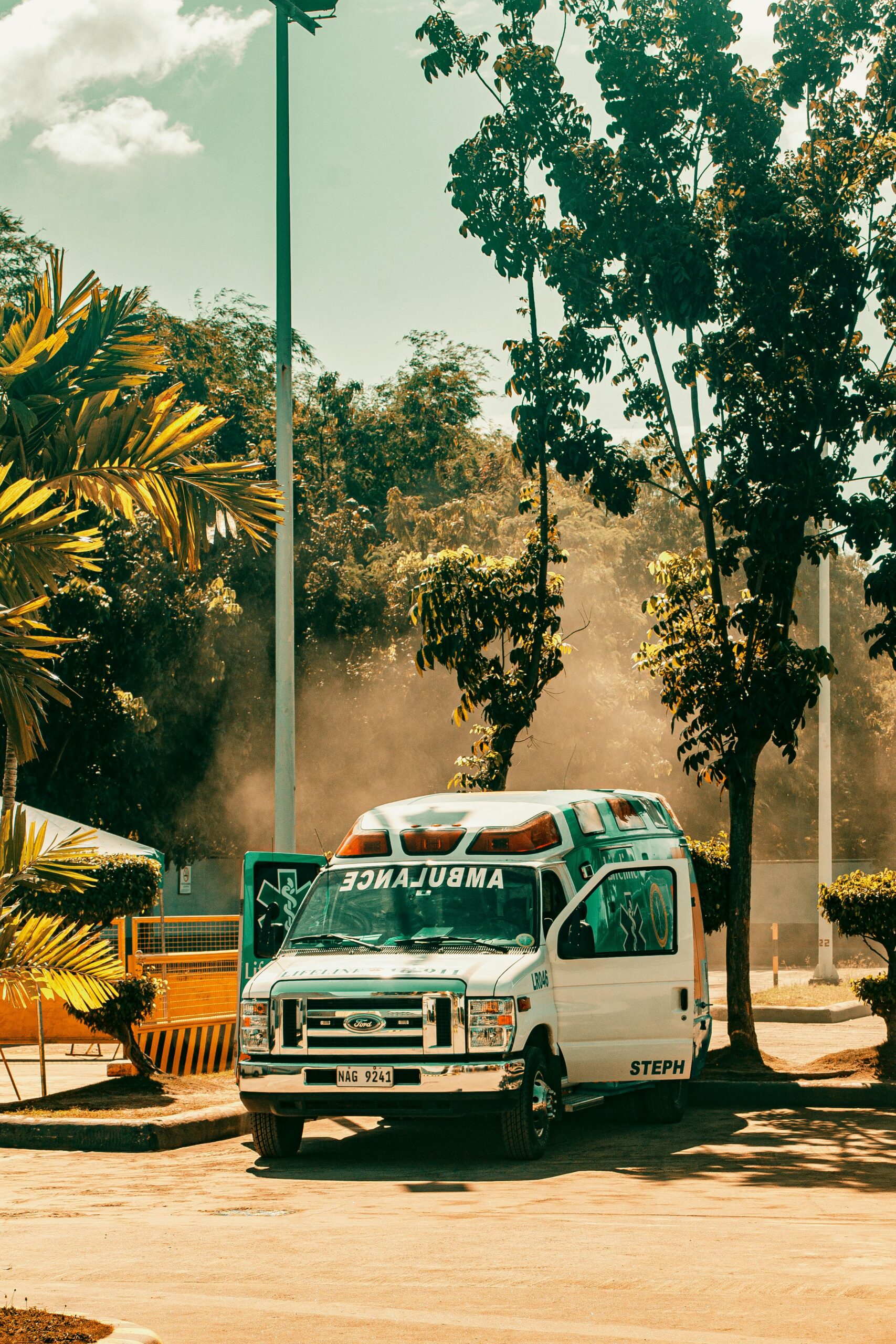 A parked ambulance in a sunlit outdoor area surrounded by trees and sunny weather.
