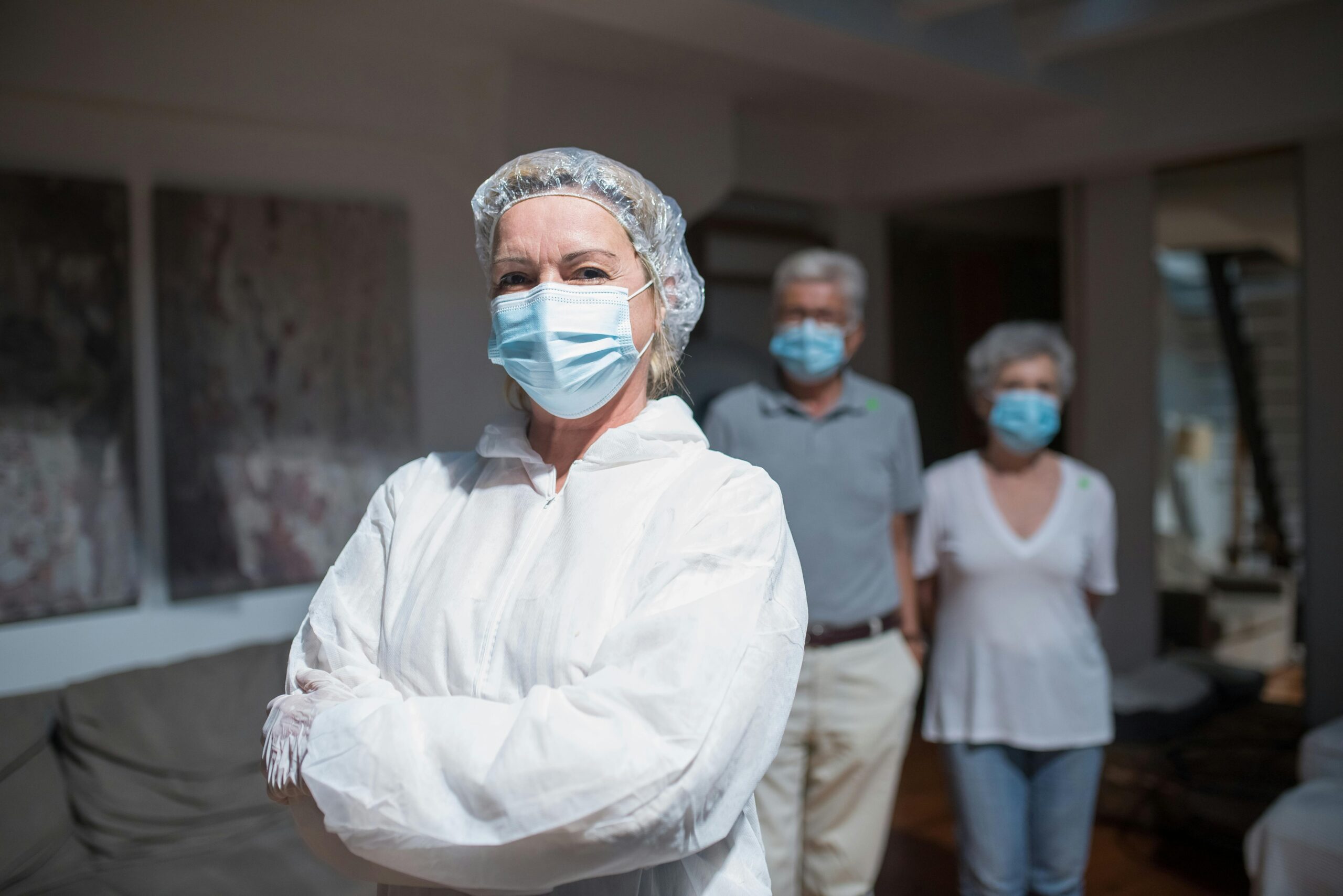 Healthcare worker in protective gear standing with an elderly couple wearing masks indoors.
